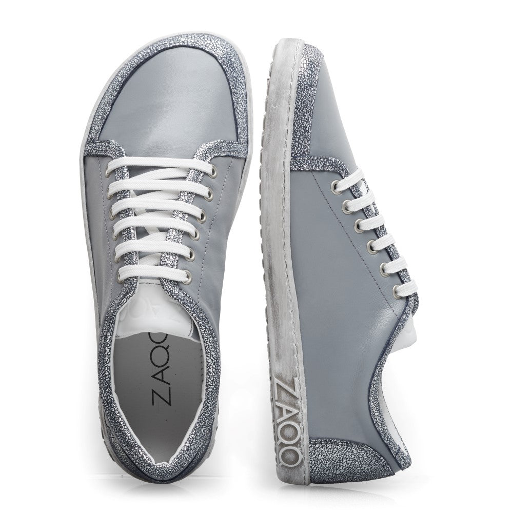 Die TIQQ Grey Silver Sneaker von ZAQQ vereinen Stil mit Komfort und sind aus hochwertigem Nappaleder gefertigt. Sie verfügen über glitzernde silberne Akzente an den Rändern und sind mit weißen Schnürsenkeln und Gummisohlen ausgestattet, die einen Komfort ähnlich einem Barfußschuh bieten. Der Markenname ZAQQ ist elegant sowohl auf der Innensohle als auch auf der Seite des Schuhs angebracht.